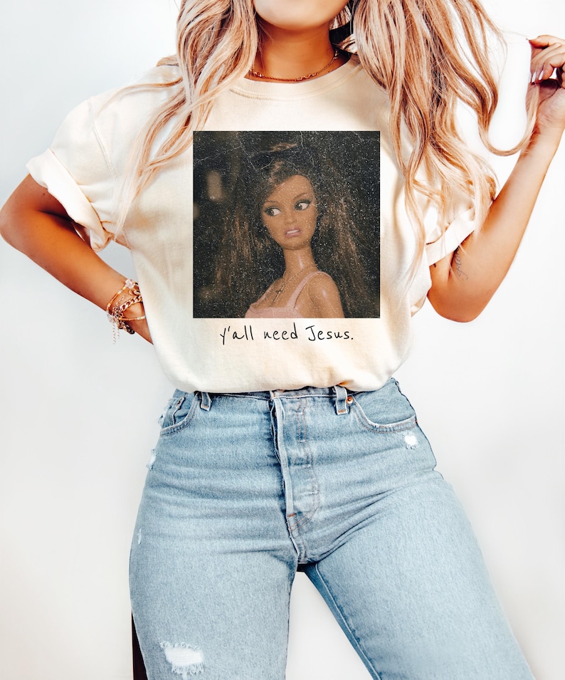 May include: A cream-colored t-shirt featuring a square graphic of a doll with long brown hair wearing a pink top. Below the graphic, the text reads, y'all need Jesus. The shirt is paired with light blue denim jeans with distressed details.