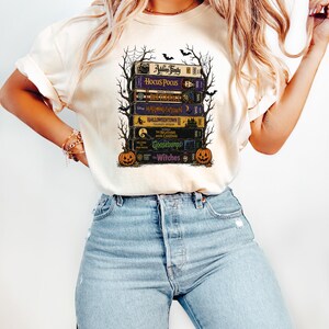 May include: A cream-colored t-shirt featuring a graphic print of a stack of vintage VHS tapes titled The Addams Family, Hocus Pocus, Beetlejuice, Halloweentown, Halloweentown II, The Nightmare Before Christmas, Goosebumps, and The Witches with pumpkins.