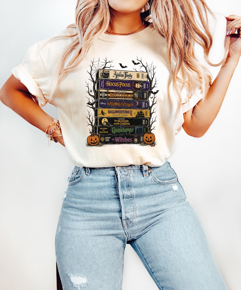 May include: A cream-colored t-shirt featuring a graphic print of a stack of vintage VHS tapes titled The Addams Family, Hocus Pocus, Beetlejuice, Halloweentown, Halloweentown II, The Nightmare Before Christmas, Goosebumps, and The Witches with pumpkins.