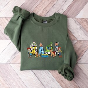 May include: An olive green sweatshirt featuring a colorful graphic of characters from a popular animated movie. The word "AM" is spelled out with the characters. The sweatshirt has long sleeves and a crew neck.