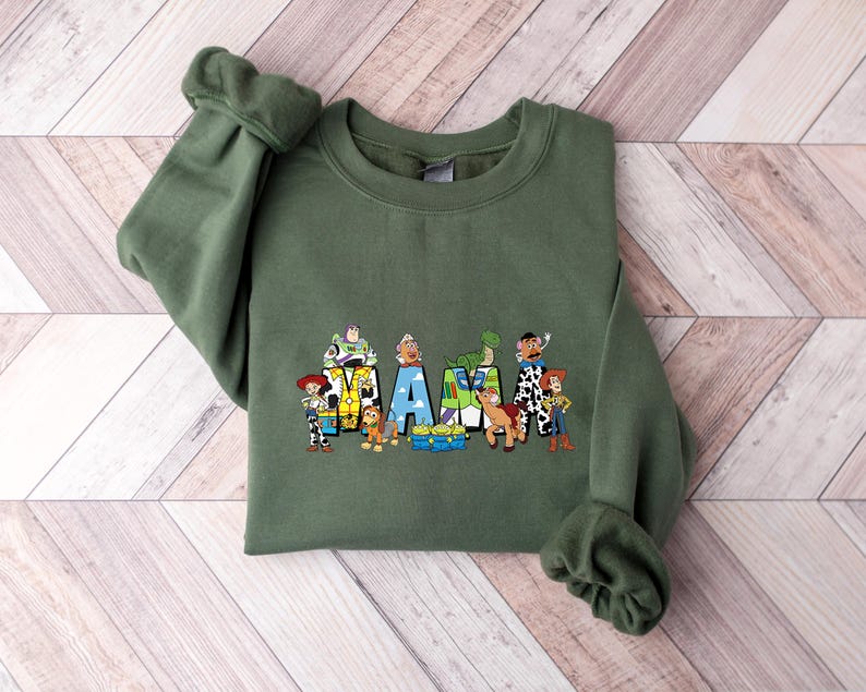 May include: An olive green sweatshirt featuring a colorful graphic of characters from a popular animated movie. The word "AM" is spelled out with the characters. The sweatshirt has long sleeves and a crew neck.
