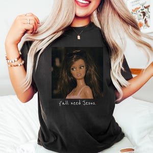 May include: A black short-sleeved t-shirt featuring a graphic print of a doll with long brown hair wearing sunglasses and a pink top. Below the doll, the text "y'all need Jesus." is printed in a simple white font on the front of the garment.