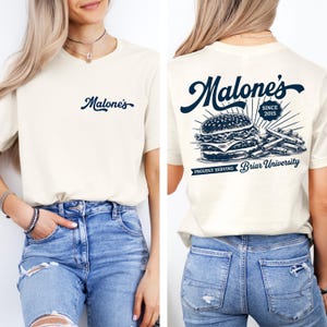 May include: Cream-colored t-shirt with the word "Malone's" in navy blue script on the front. The back features a navy blue graphic of a burger and fries, with the text "Malone's Since 2015" and "Proudly Serving Briar University."