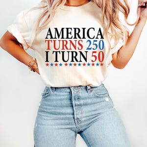 May include: Cream-colored t-shirt with the text "AMERICA TURNS 250 I TURN 50" in black, red, and blue. The text is above a row of red and blue stars. The person is wearing light blue jeans.