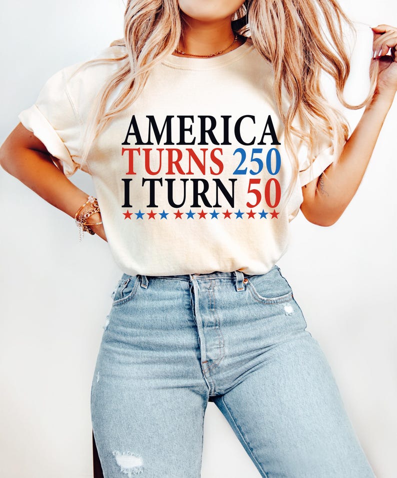 May include: Cream-colored t-shirt with the text "AMERICA TURNS 250 I TURN 50" in black, red, and blue. The text is above a row of red and blue stars. The person is wearing light blue jeans.