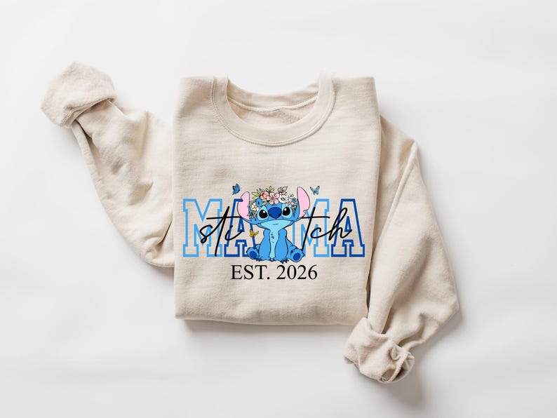 Puede incluir: Sudadera beige con un personaje de dibujos animados azul con una corona de flores y el texto "Stitch Mama EST. 2026". Las mangas est&aacute;n enrolladas. El dise&ntilde;o es en tonos azules.