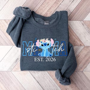Puede incluir: Una sudadera gris oscuro con un personaje de dibujos animados azul con una corona de flores y el texto "Mama Stitch EST. 2026" en azul y blanco. La sudadera est&aacute; doblada sobre una superficie de madera.