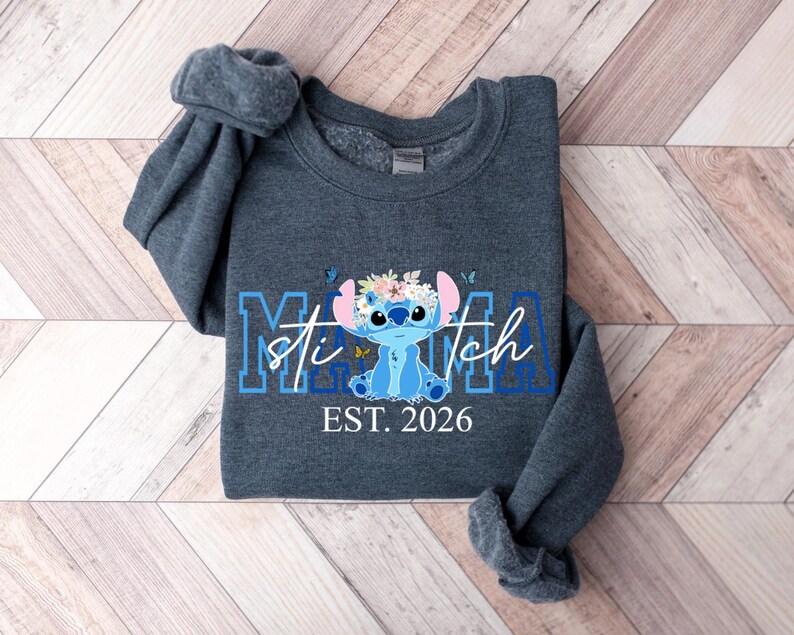 Puede incluir: Una sudadera gris oscuro con un personaje de dibujos animados azul con una corona de flores y el texto "Mama Stitch EST. 2026" en azul y blanco. La sudadera est&aacute; doblada sobre una superficie de madera.
