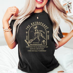 May include: Black t-shirt with a gold graphic featuring an astronaut, sun, and text that reads "THE ASTROPHAGE". The text also includes "ENERGY DEVOURER OF THE STARS" and a quote about organisms that eat stars.