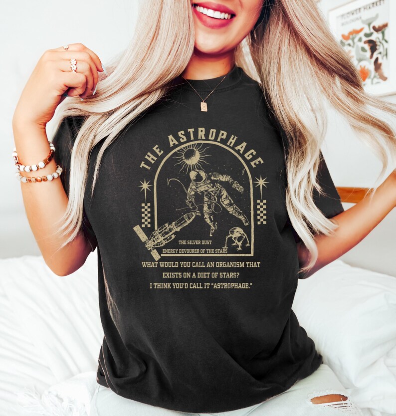 May include: Black t-shirt with a gold graphic featuring an astronaut, sun, and text that reads "THE ASTROPHAGE". The text also includes "ENERGY DEVOURER OF THE STARS" and a quote about organisms that eat stars.