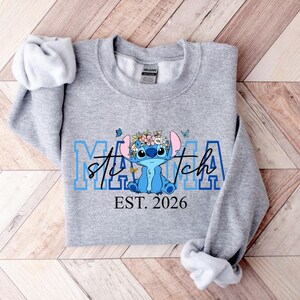 Puede incluir: Una sudadera gris claro con un personaje de dibujos animados azul con una corona de flores. La palabra "Mama" est&aacute; en letras azules, con "Stitch" en cursiva. Debajo, el texto dice "EST. 2026".