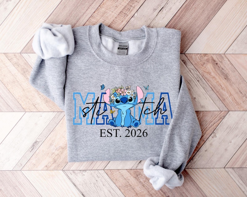 Puede incluir: Una sudadera gris claro con un personaje de dibujos animados azul con una corona de flores. La palabra "Mama" est&aacute; en letras azules, con "Stitch" en cursiva. Debajo, el texto dice "EST. 2026".