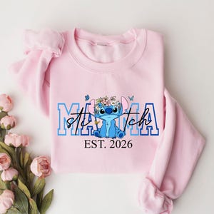 Puede incluir: Una sudadera rosa claro con las palabras "Mama Stitch" en letras azules y blancas, con un personaje de dibujos animados con una corona de flores. El texto "EST. 2026" est&aacute; debajo.