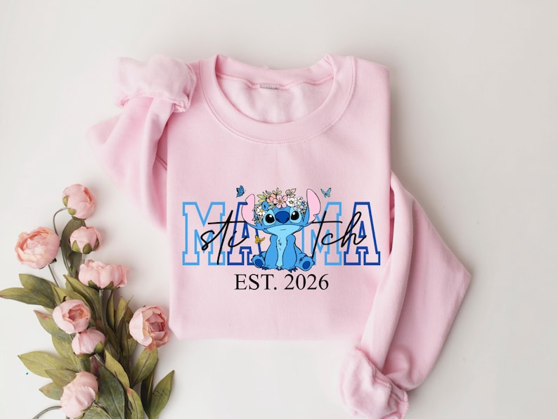 Puede incluir: Una sudadera rosa claro con las palabras "Mama Stitch" en letras azules y blancas, con un personaje de dibujos animados con una corona de flores. El texto "EST. 2026" est&aacute; debajo.