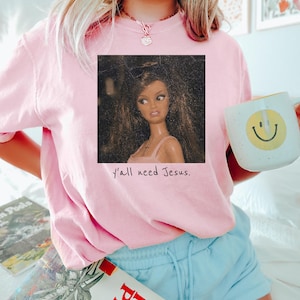 May include: A light pink short-sleeve t-shirt featuring a square graphic of a doll with long brown hair and a pink top. Below the graphic, the text reads y'all need Jesus. The shirt is paired with light blue shorts and a white mug with a yellow smiley face.