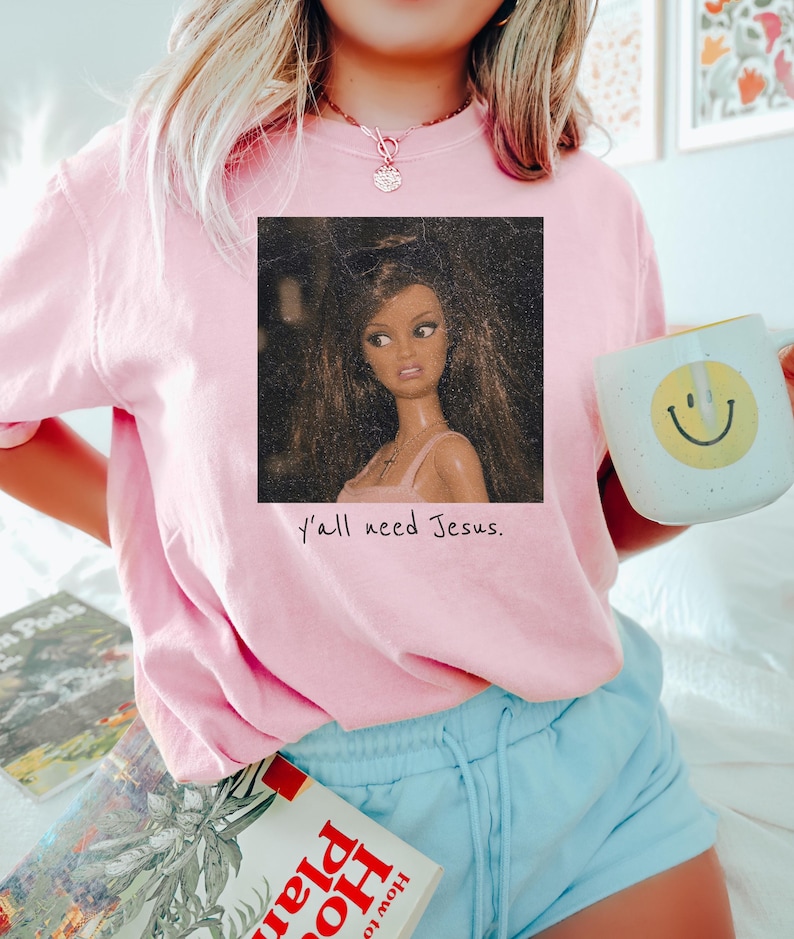May include: A light pink short-sleeve t-shirt featuring a square graphic of a doll with long brown hair and a pink top. Below the graphic, the text reads y'all need Jesus. The shirt is paired with light blue shorts and a white mug with a yellow smiley face.