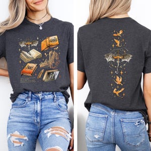May include: Dark gray t-shirt featuring a fantasy book design on the front, with open books, a castle, and a dragon. The back of the shirt has a dragon and star design. The shirt is paired with blue jeans.