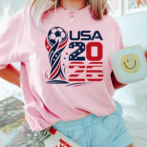 Op de afbeelding: Een lichtroze t-shirt met een grafisch ontwerp van een voetbal bovenop een trofee, de tekst USA 2026 en rode, witte en blauwe sterren- en strepenpatronen. Het ontwerp is gecentreerd op de borst van het kledingstuk met korte mouwen.