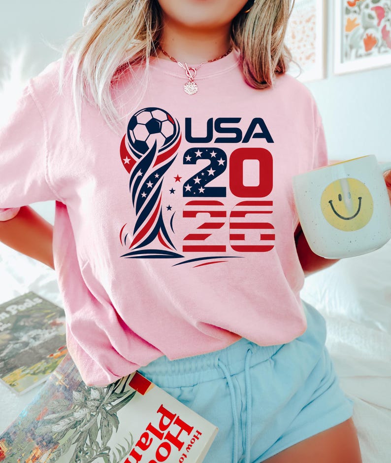 Op de afbeelding: Een lichtroze t-shirt met een grafisch ontwerp van een voetbal bovenop een trofee, de tekst USA 2026 en rode, witte en blauwe sterren- en strepenpatronen. Het ontwerp is gecentreerd op de borst van het kledingstuk met korte mouwen.