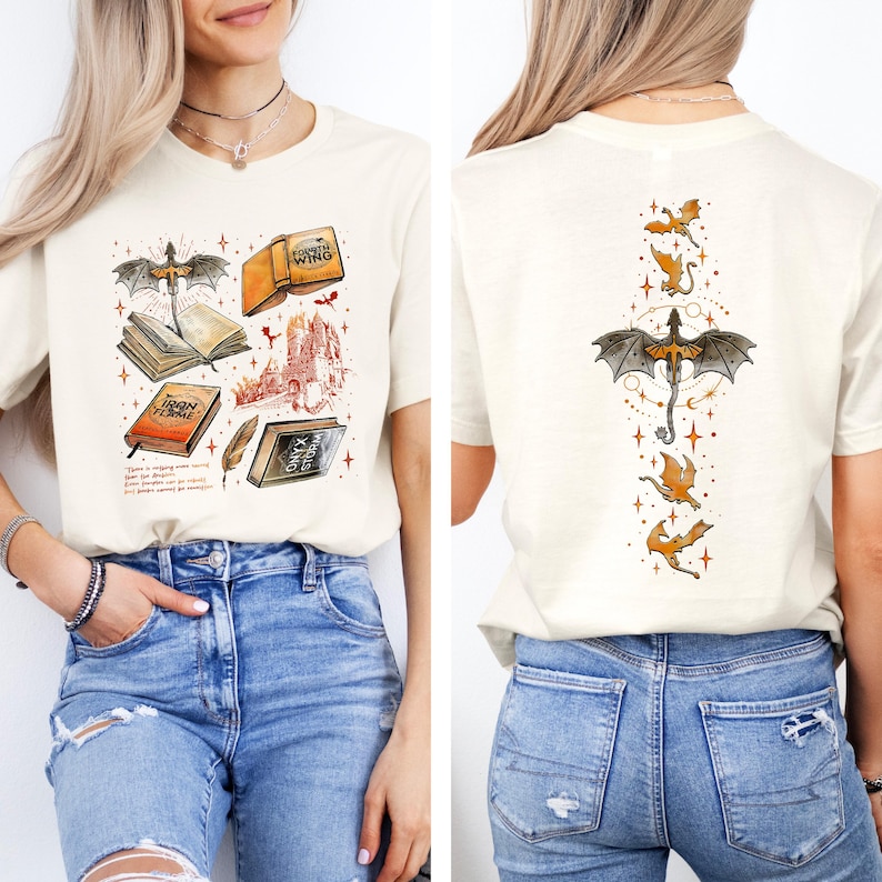 May include: Cream-colored t-shirt featuring a fantasy-themed illustration on the front, including books, a castle, and a dragon. The back of the shirt displays a dragon and several smaller dragons. The design incorporates orange, black, and brown elements.