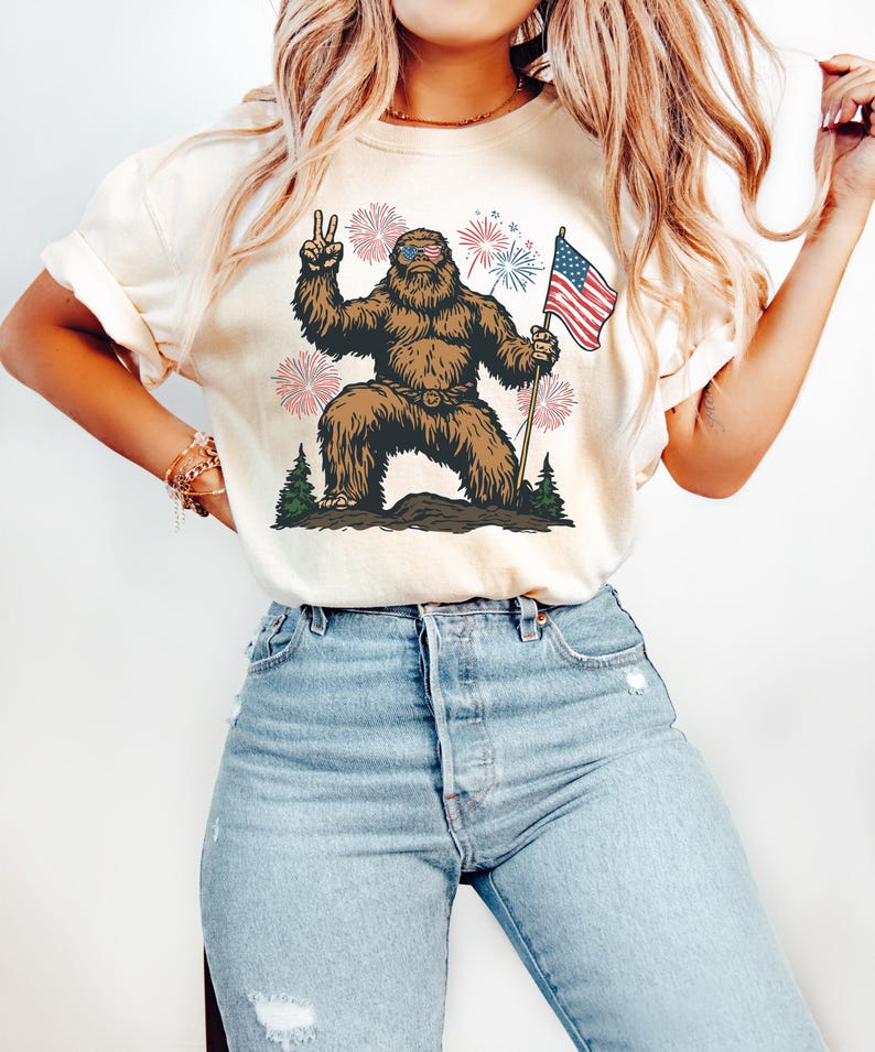 Pu&ograve; includere: Una t-shirt color crema con la grafica di un Bigfoot marrone che indossa occhiali da sole con il motivo della bandiera americana, fa il segno di pace e tiene in mano una bandiera americana. Il design include fuochi d'artificio e pini.