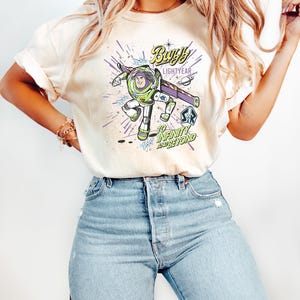May include: Cream-colored t-shirt featuring a graphic of Buzz Lightyear in a dynamic pose, with the text "Buzz Lightyear" and "To Infinity and Beyond" in a stylized font. The shirt is paired with light blue jeans.