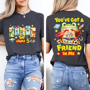 May include: A dark gray t-shirt featuring Toy Story graphics. The front displays characters in rectangular frames with the text "TOY STORY 5". The back shows characters watching a screen with the text "YOU'VE GOT A FRIEND IN ME".