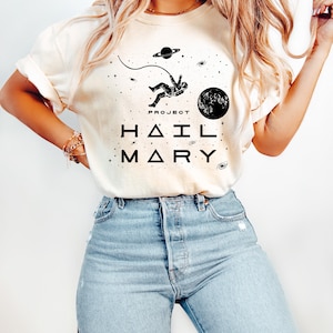 May include: A cream-coloured t-shirt with a black graphic of an astronaut in space, a planet, and stars. The text "PROJECT" is above the astronaut, and "HAIL MARY" is printed below. The shirt is worn with light blue jeans.
