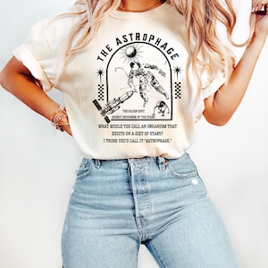 May include: Cream-colored t-shirt featuring a black and white graphic of an astronaut, a sun, and a spaceship, with the text "THE ASTROPHAGE." The shirt also includes the text "THE SILVER DUST ENERGY DEVOURER OF THE STARS."