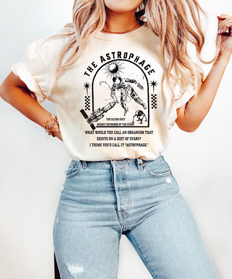 May include: Cream-colored t-shirt featuring a black and white graphic of an astronaut, a sun, and a spaceship, with the text "THE ASTROPHAGE." The shirt also includes the text "THE SILVER DUST ENERGY DEVOURER OF THE STARS."