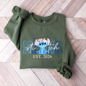 Puede incluir: Una sudadera verde oliva con un dise&ntilde;o azul y blanco de Stitch, la palabra "Mama" y "Stitch" en azul, y el texto "EST. 2026" en blanco. El dise&ntilde;o incluye mariposas y detalles florales.