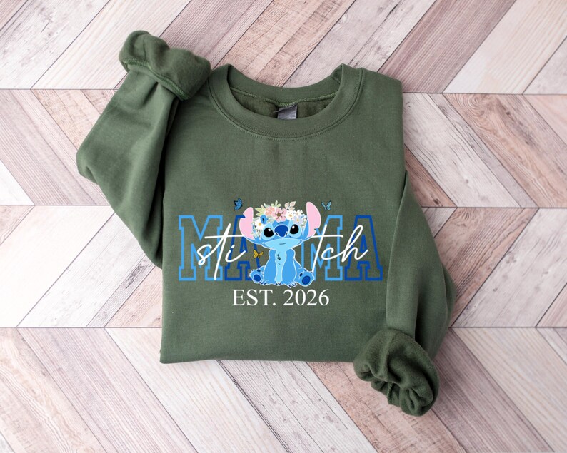 Puede incluir: Una sudadera verde oliva con un dise&ntilde;o azul y blanco de Stitch, la palabra "Mama" y "Stitch" en azul, y el texto "EST. 2026" en blanco. El dise&ntilde;o incluye mariposas y detalles florales.