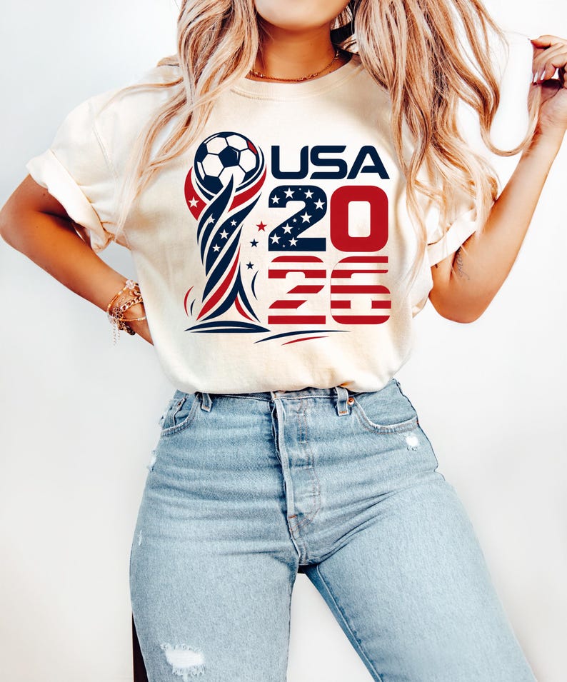 Op de afbeelding: Een cr&egrave;mekleurig T-shirt met korte mouwen en een grafische print van een voetbal boven op een trofee, met de tekst USA 2026. Het ontwerp bevat rode, witte en blauwe kleuren met sterpatronen, gecombineerd met een lichtblauwe spijkerbroek.