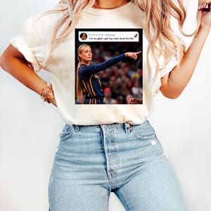 May include: A cream-colored short-sleeve t-shirt featuring a graphic print of a social media post. The graphic shows a blonde athlete in a dark blue jersey pointing, with the text "SOPHIE Following I'm so glad I got my nails done for this" and a TikTok logo.