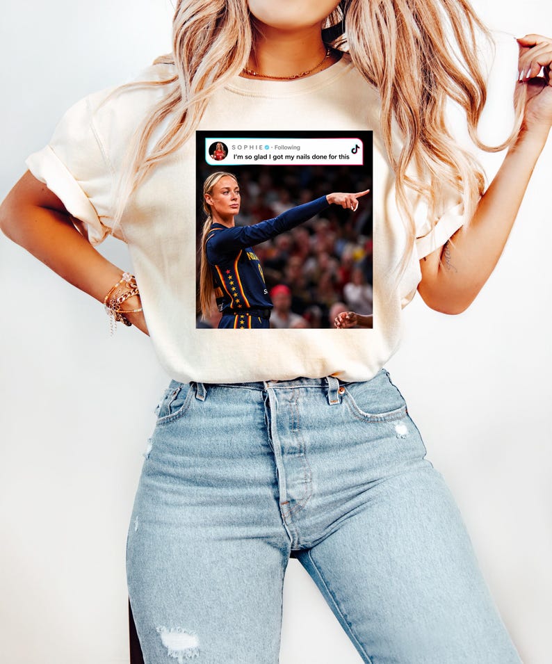 May include: A cream-colored short-sleeve t-shirt featuring a graphic print of a social media post. The graphic shows a blonde athlete in a dark blue jersey pointing, with the text "SOPHIE Following I'm so glad I got my nails done for this" and a TikTok logo.