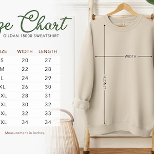 May include: A beige Gildan 18000 sweatshirt hangs on a wooden rack. The image includes a size chart with measurements in inches: S (20" width, 27" length), M (22" width, 28" length), L (24" width, 29" length), XL (26" width, 30" length), 2XL (28" width, 31" length), 3XL (30" width, 32" length), 4XL (32" width, 33" length), and 5XL (34" width, 34" length).