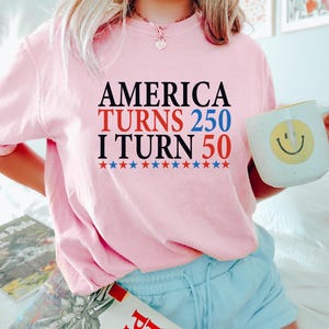 May include: Pink sweatshirt with the text "AMERICA TURNS 250 I TURN 50" in black, red, and blue. Below the text are red, white, and blue stars. A white mug with a yellow smiley face is held in the hand.