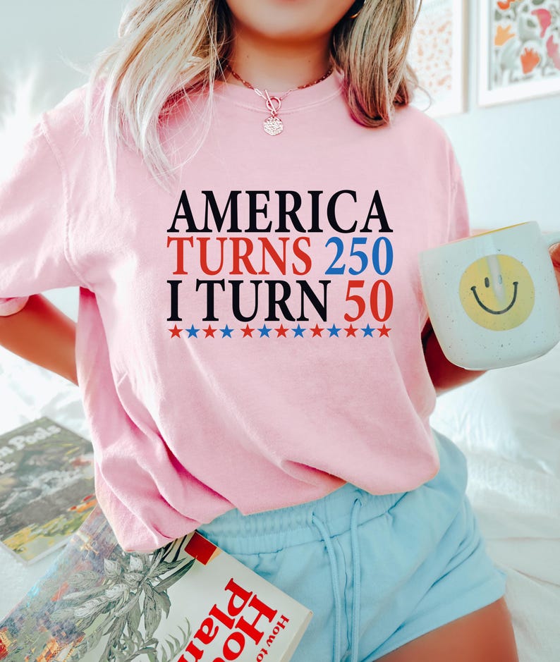 May include: Pink sweatshirt with the text "AMERICA TURNS 250 I TURN 50" in black, red, and blue. Below the text are red, white, and blue stars. A white mug with a yellow smiley face is held in the hand.