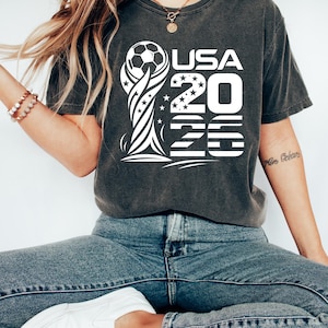 Op de afbeelding: Een antracietgrijs T-shirt met korte mouwen en een witte grafische print van een voetbal op een trofeesilhouet, met de tekst USA 2026 in het ontwerp verwerkt. De afbeelding bevat steraccenten en horizontale lijnpatronen.