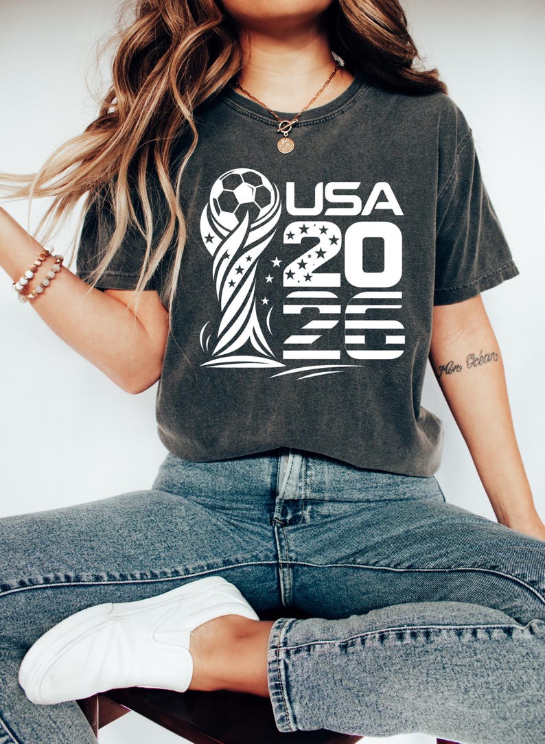 Op de afbeelding: Een antracietgrijs T-shirt met korte mouwen en een witte grafische print van een voetbal op een trofeesilhouet, met de tekst USA 2026 in het ontwerp verwerkt. De afbeelding bevat steraccenten en horizontale lijnpatronen.
