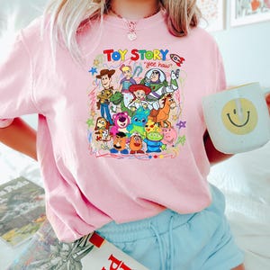 May include: Pink crew neck t-shirt featuring a colorful graphic of Toy Story characters, including Woody, Buzz Lightyear, and others. The text "Toy Story" and "yee haw" are also visible. The shirt is worn by a person holding a mug.