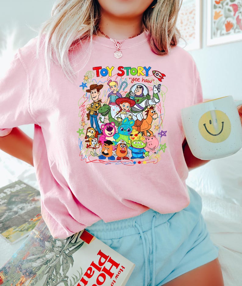 May include: Pink crew neck t-shirt featuring a colorful graphic of Toy Story characters, including Woody, Buzz Lightyear, and others. The text "Toy Story" and "yee haw" are also visible. The shirt is worn by a person holding a mug.