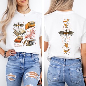 May include: White t-shirt featuring a fantasy-themed graphic on the front and back. The front design includes books, a dragon, and a castle. The back features a dragon and smaller flying dragons. The shirt is paired with blue jeans.