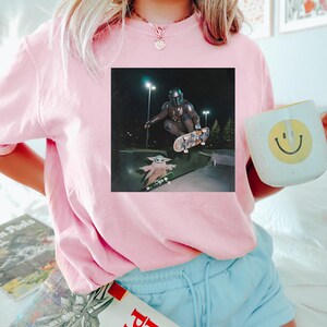 Puede incluir: Una sudadera rosa claro con un gr&aacute;fico cuadrado de The Mandalorian y Grogu patinando de noche. La persona de la imagen sostiene una taza blanca con una carita sonriente amarilla. La persona tambi&eacute;n lleva pantalones cortos de color azul claro.