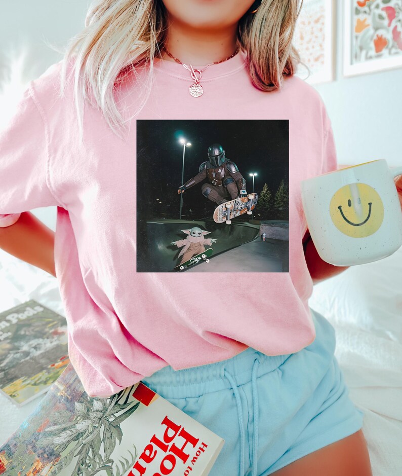 Puede incluir: Una sudadera rosa claro con un gr&aacute;fico cuadrado de The Mandalorian y Grogu patinando de noche. La persona de la imagen sostiene una taza blanca con una carita sonriente amarilla. La persona tambi&eacute;n lleva pantalones cortos de color azul claro.