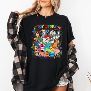 May include: Black t-shirt featuring a colorful graphic of characters from the animated film "Toy Story." The design includes Woody, Buzz Lightyear, and other beloved characters, with the words "Toy Story" in a vibrant, multi-colored font.