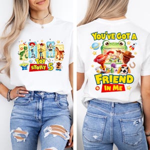 May include: A white t-shirt featuring Toy Story graphics. The front displays characters like Woody, Buzz Lightyear, and Rex with the text "TOY STORY 5". The back shows a frog-themed screen with characters and the text "YOU'VE GOT A FRIEND IN ME".