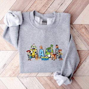 May include: A gray sweatshirt with the word "MAMA" in colorful block letters, each letter featuring characters from a popular animated film. The sweatshirt has long sleeves and a crew neck. The design includes characters like Woody, Buzz Lightyear, and Rex.