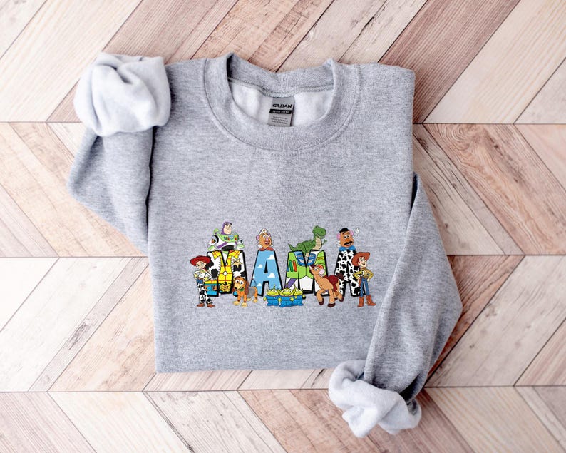 May include: A gray sweatshirt with the word "MAMA" in colorful block letters, each letter featuring characters from a popular animated film. The sweatshirt has long sleeves and a crew neck. The design includes characters like Woody, Buzz Lightyear, and Rex.