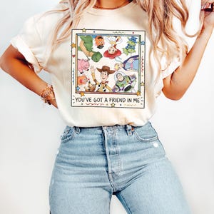 Op de afbeelding: Een cr&egrave;mekleurig T-shirt met korte mouwen en een vierkante grafische print van verschillende geanimeerde Toy Story-personages in een cirkel. Onder de personages staat de tekst "YOU'VE GOT A FRIEND IN ME" in een eenvoudig, donker lettertype.